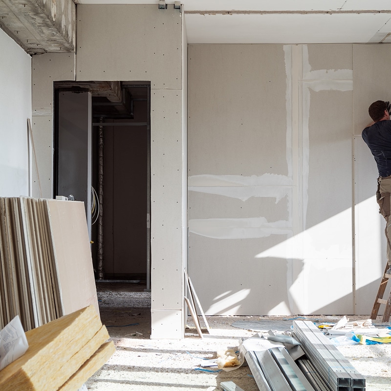 Ouvrier installant des plaques de plâtre (placoplatre) sur un mur en construction, matériaux de finition empilés au sol.