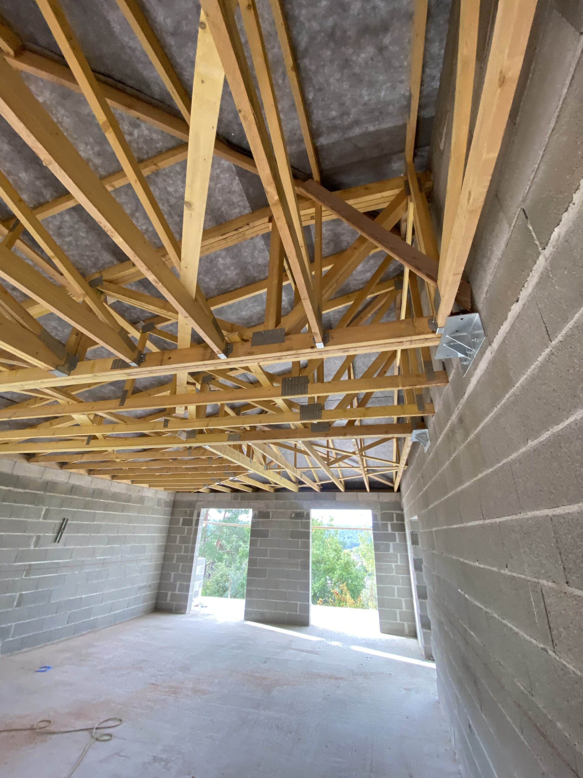 Intérieur d'une maison en construction, murs en parpaings gris, charpente apparente en bois et deux ouvertures donnant sur la verdure.