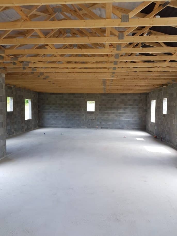 Intérieur brut d'un bâtiment avec murs en parpaings, plancher béton neuf et charpente apparente en bois clair et métal.