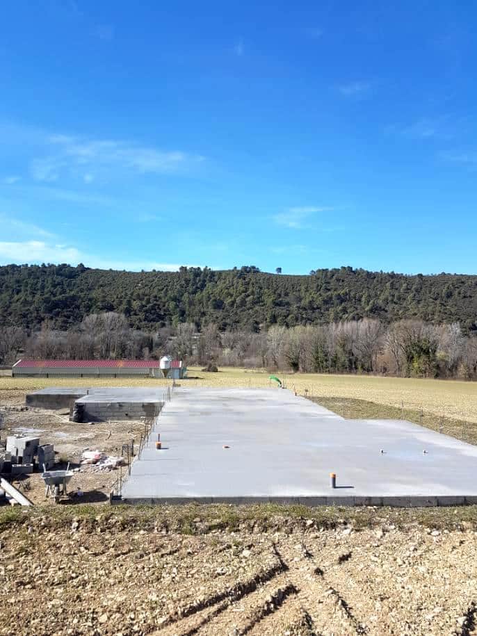 Dalle de béton fraîchement coulée pour une construction, avec des parpaings et une brouette à gauche, sur fond de campagne et de colline boisée.