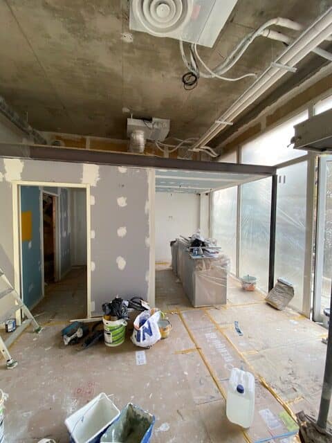 Chantier intérieur de rénovation. Plafond béton, tuyauterie exposée, mur en cours de jointoiement. Comptoir emballé et REHAU au sol.