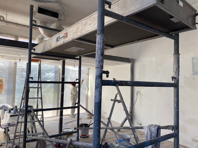 Intérieur en rénovation. Échafaudage Comabi au premier plan. Un ouvrier peintre applique la peinture sur un mur de fond. Matériel et échelles.