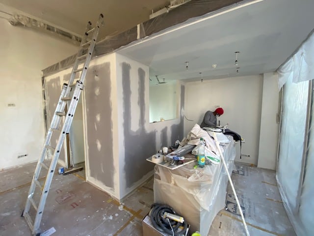 Chantier de rénovation intérieure avec plaques de plâtre et enduit. Un ouvrier ponce la zone cuisine. Échelle en aluminium.