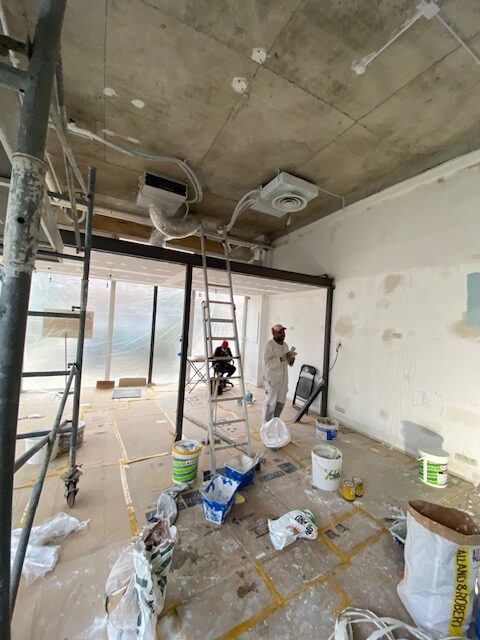 Chantier de rénovation : ouvriers préparant les murs et plafond en béton brut. Échelle, échafaudage et pots de peinture au sol.