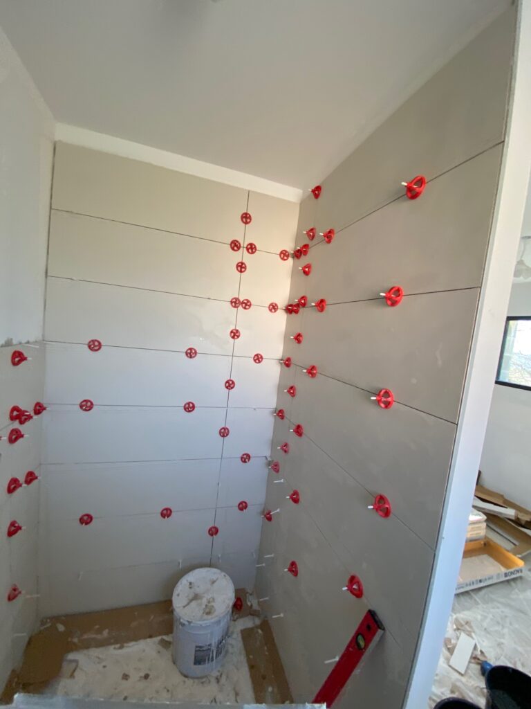 Pose de carrelage mural grand format avec croisillons autonivelants rouges dans une salle de bain en rénovation.