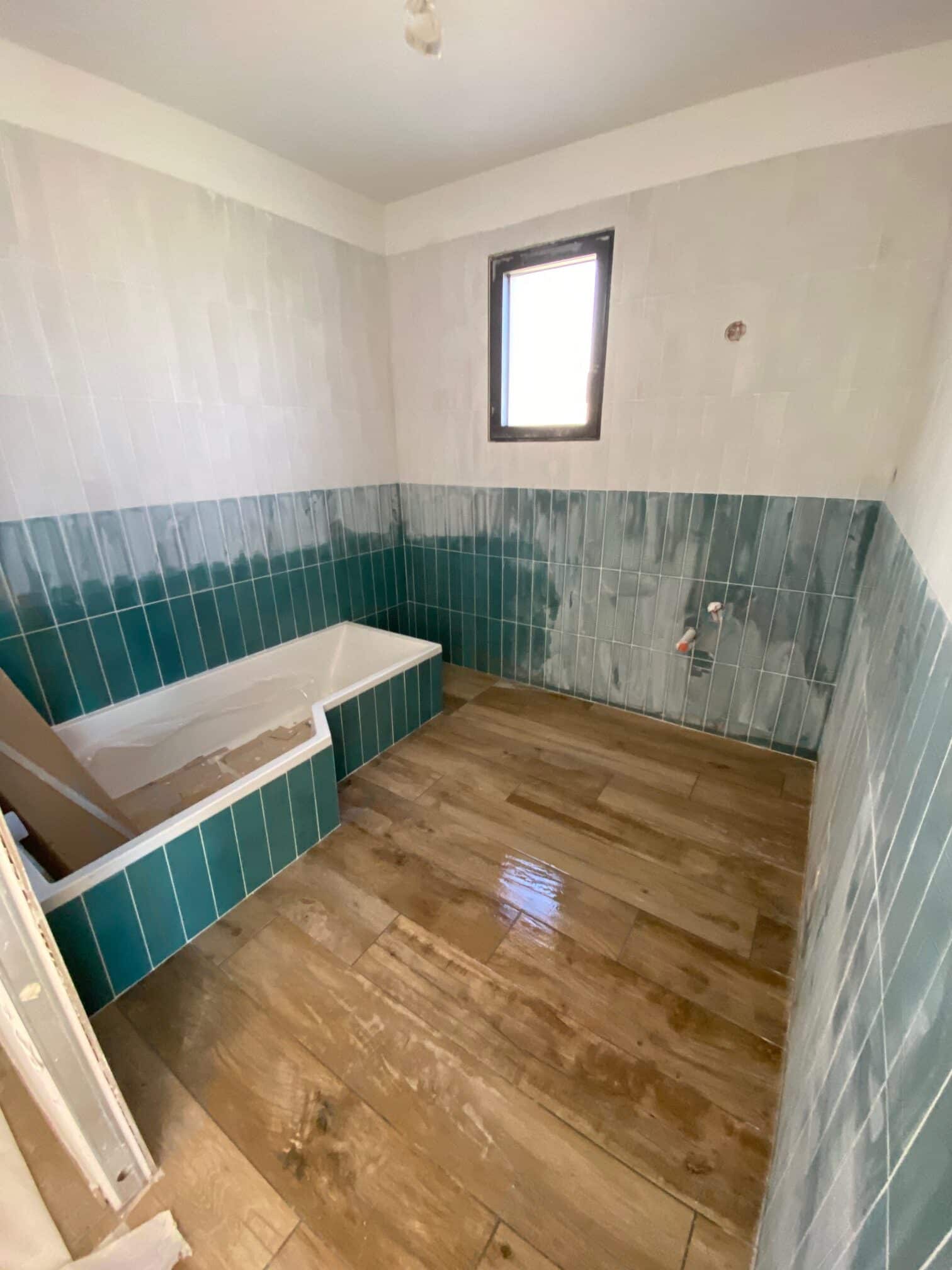 Rénovation de salle de bain avec baignoire carrelée en vert sarcelle et carrelage de sol imitation bois clair.