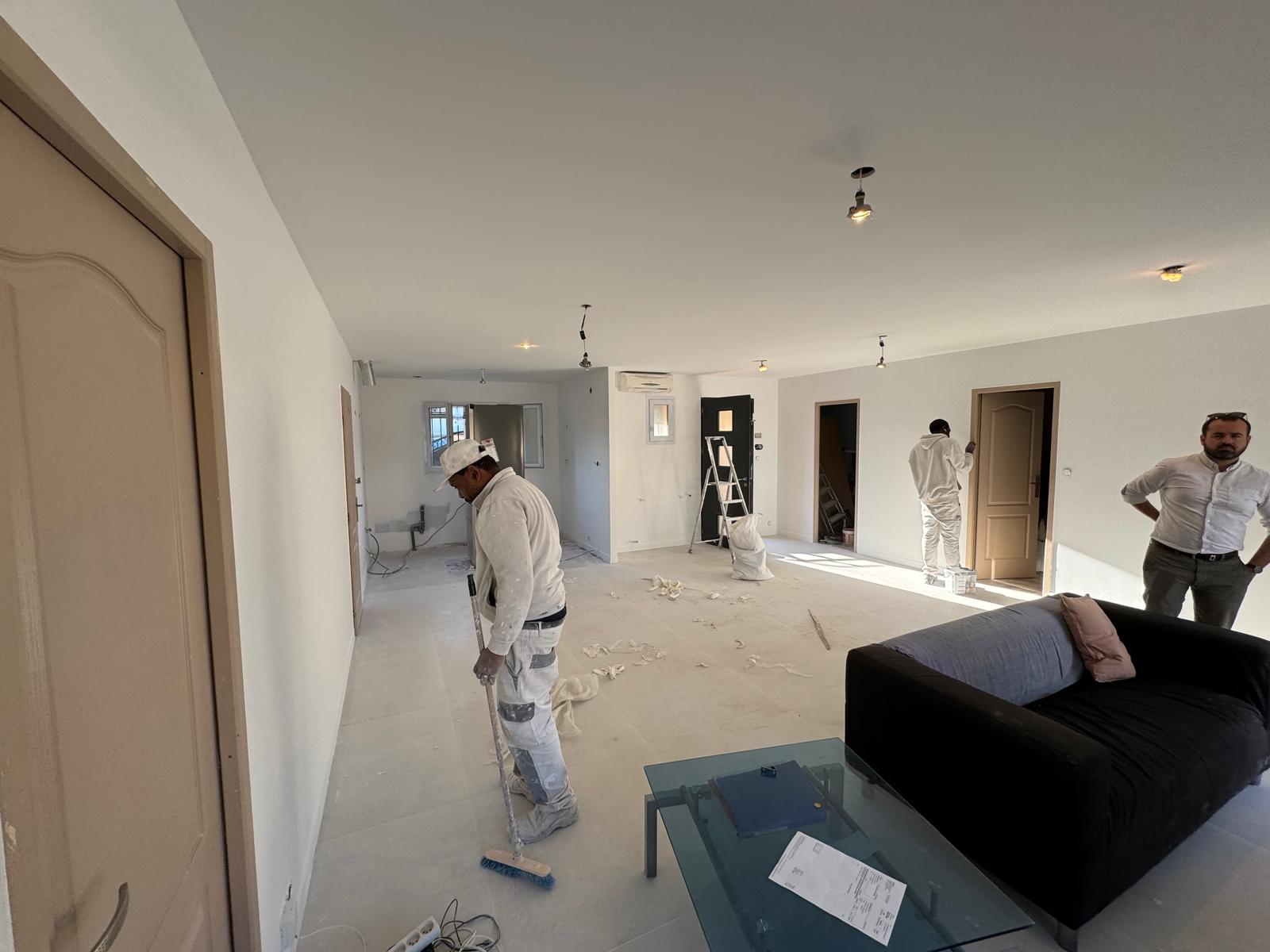 Peintres en plein travail de rénovation dans un salon blanc ; un ouvrier balaie les débris au sol.