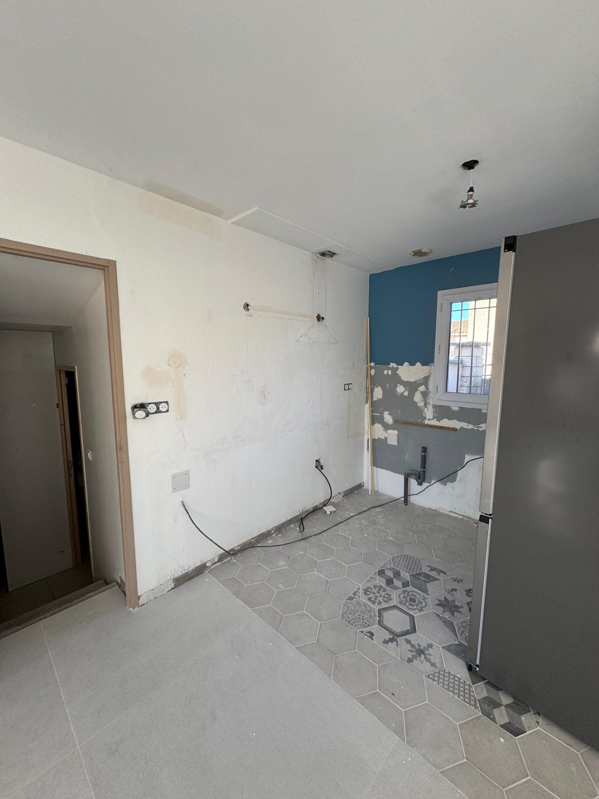 Cuisine en travaux avec murs à nu, carrelage hexagonal gris et plomberie apparente sous une petite fenêtre.
