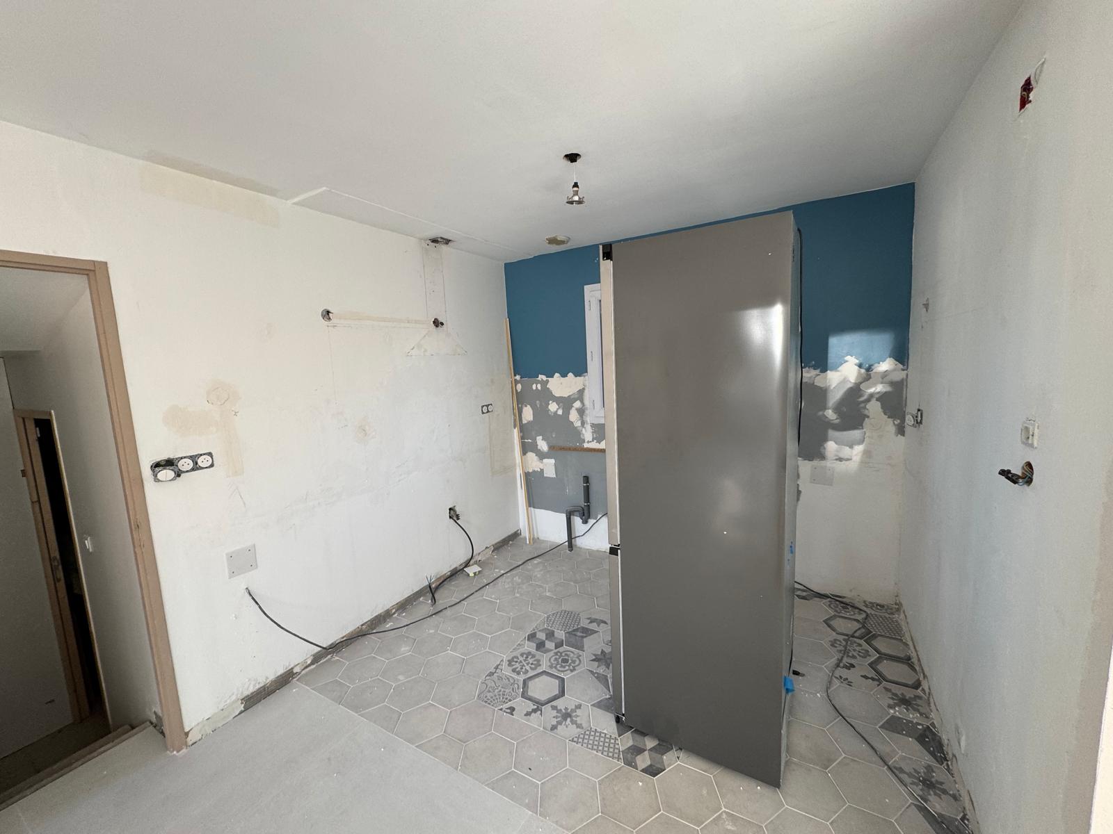 Rénovation cuisine : carrelage hexagonal gris, mur bleu et frigo inox en cours d'installation.