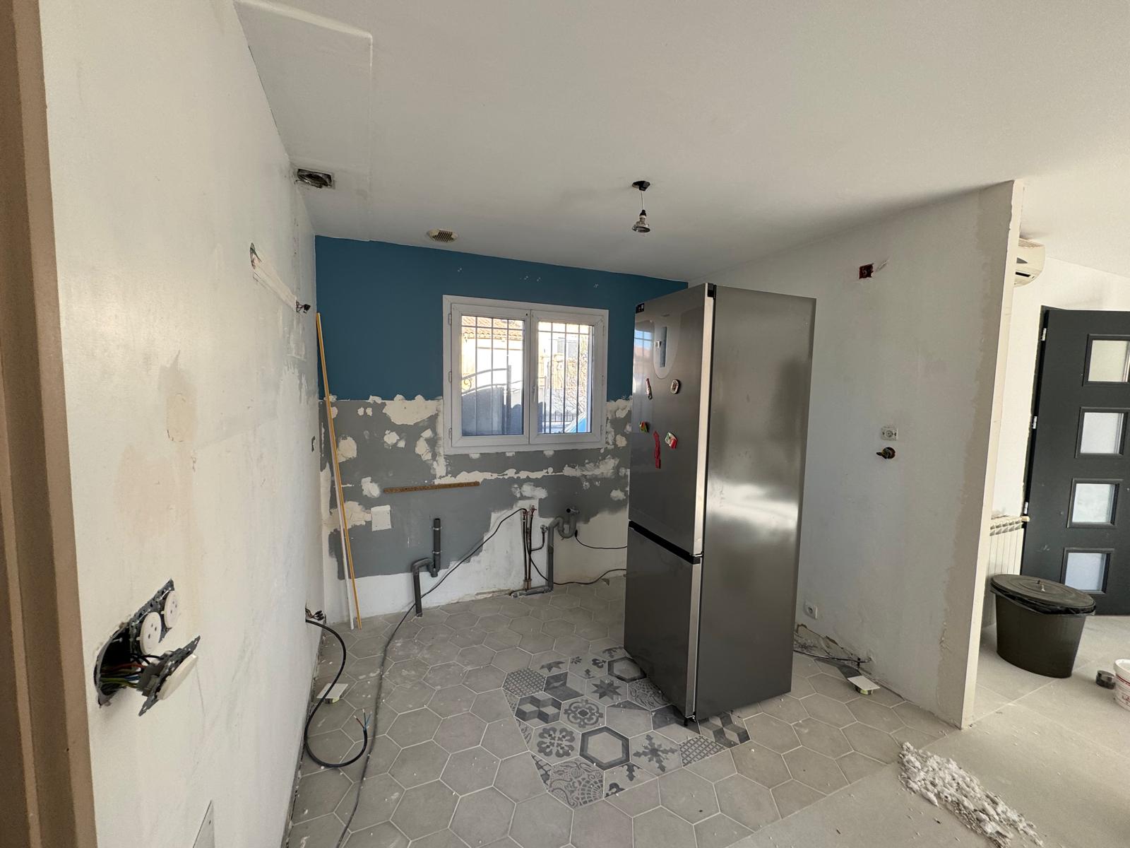 Cuisine en rénovation : murs bruts, tuyaux apparents, carrelage hexagonal gris et réfrigérateur inox.