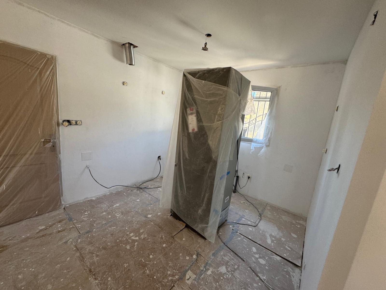 Cuisine en rénovation avec murs blancs, frigo sous plastique et sol protégé. Fils électriques et conduits visibles.