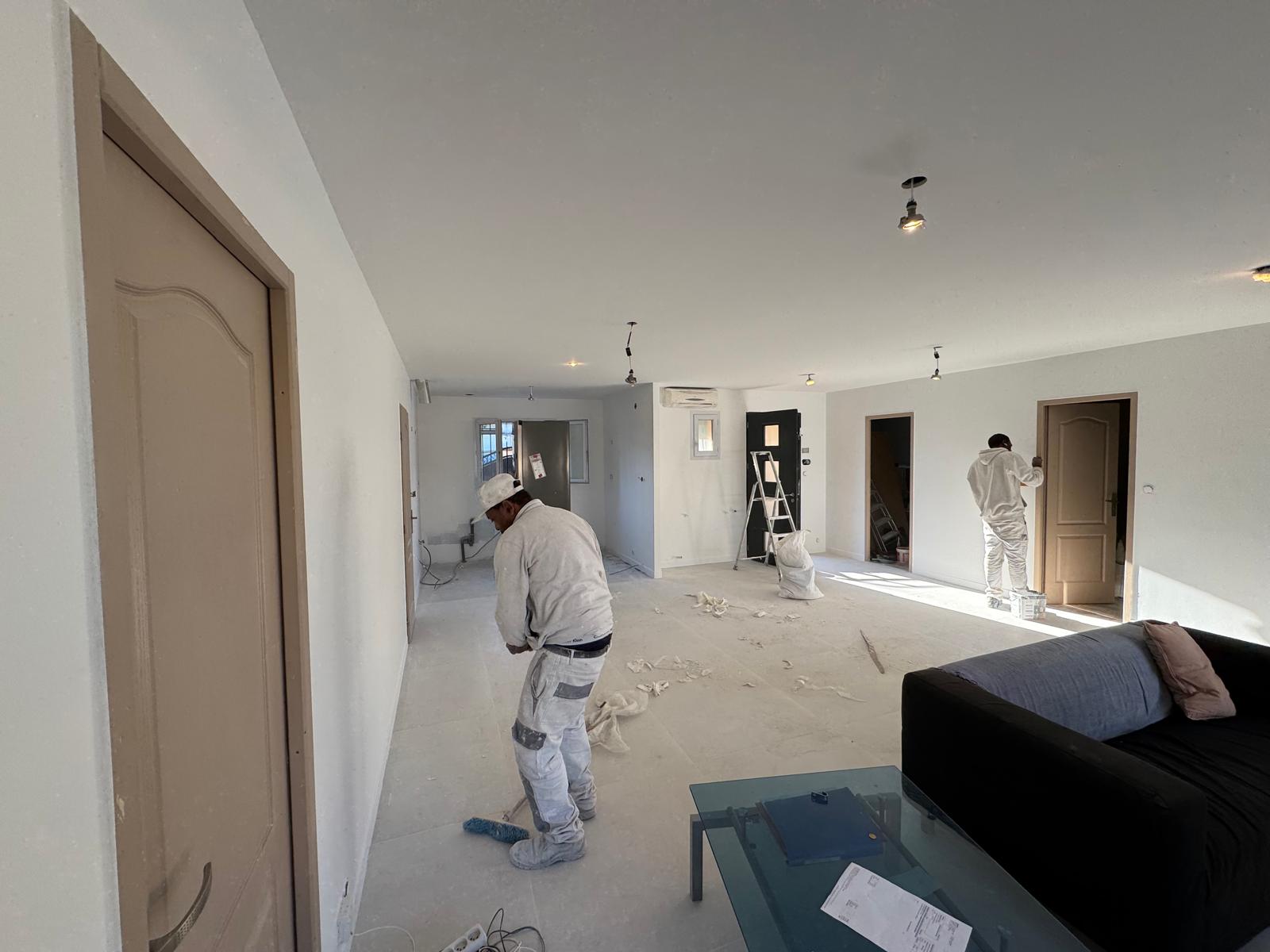 Deux peintres en tenue blanche rénovent un salon lumineux avec canapé noir et table basse.