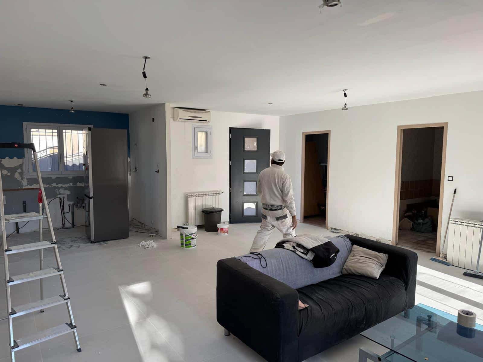 Peintre rénovant un salon avec canapé noir, échelle et murs en cours de peinture blanche.
