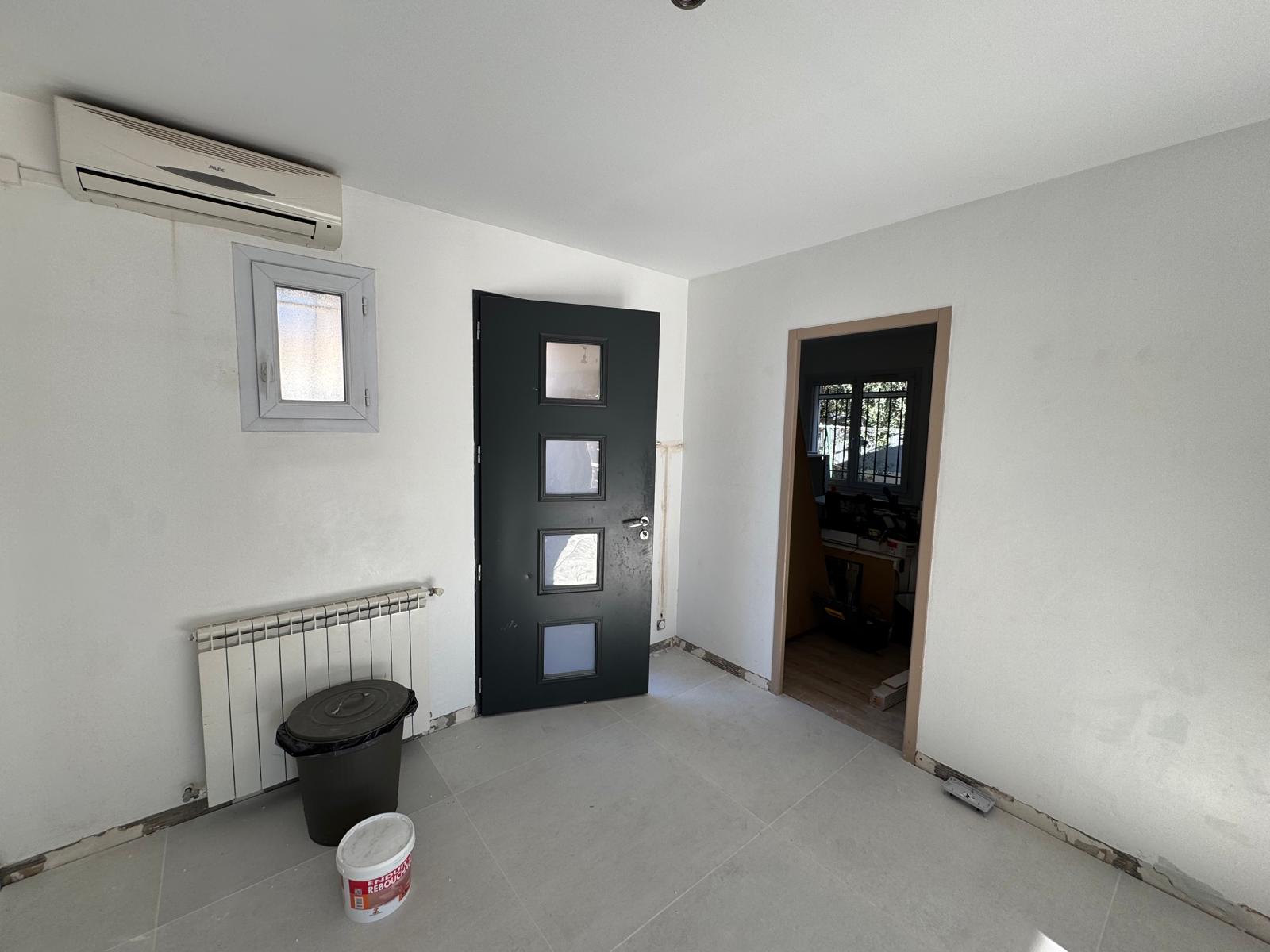 Pièce en rénovation avec porte noire vitrée, radiateur blanc et climatisation sur murs blancs.