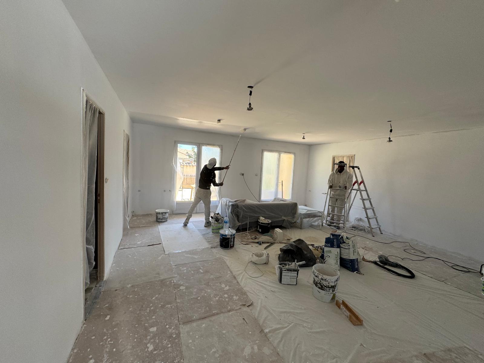 Deux peintres rénovent un salon blanc avec protection au sol et matériel de peinture.