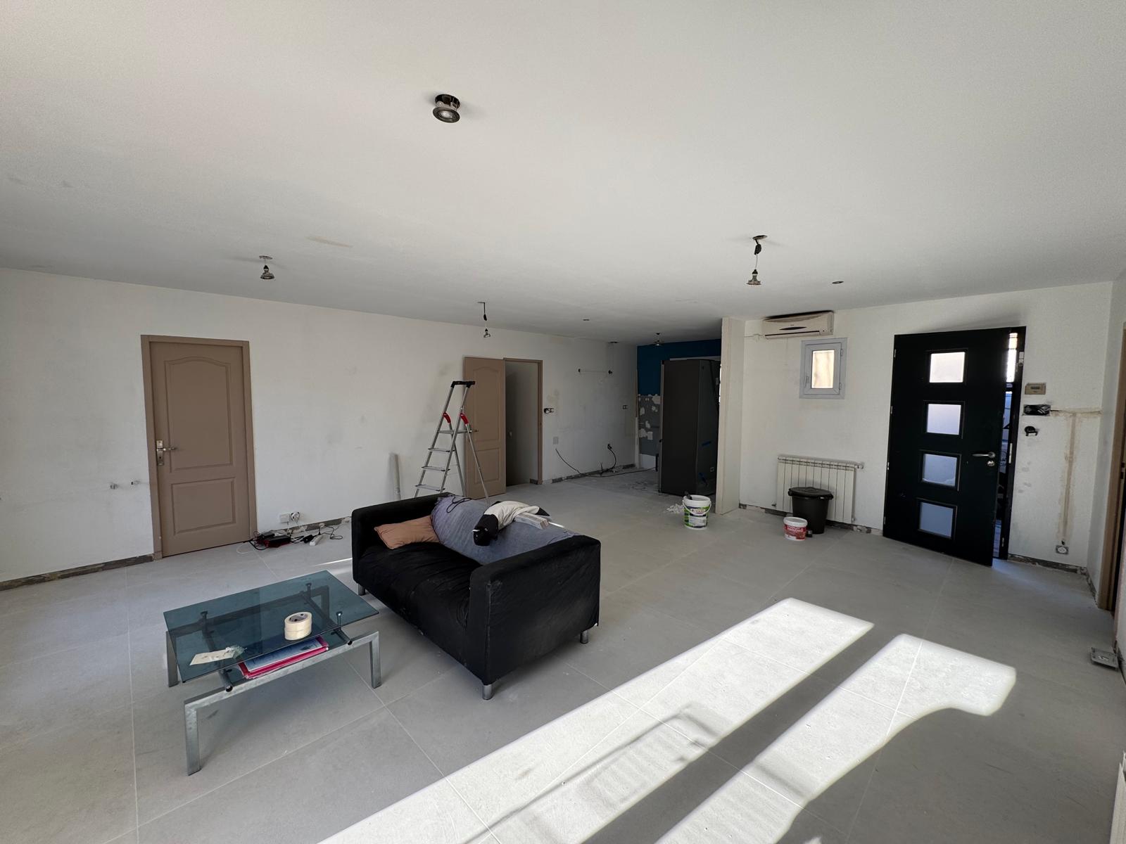 Salon en rénovation avec canapé noir et échelle de chantier sur carrelage gris clair.