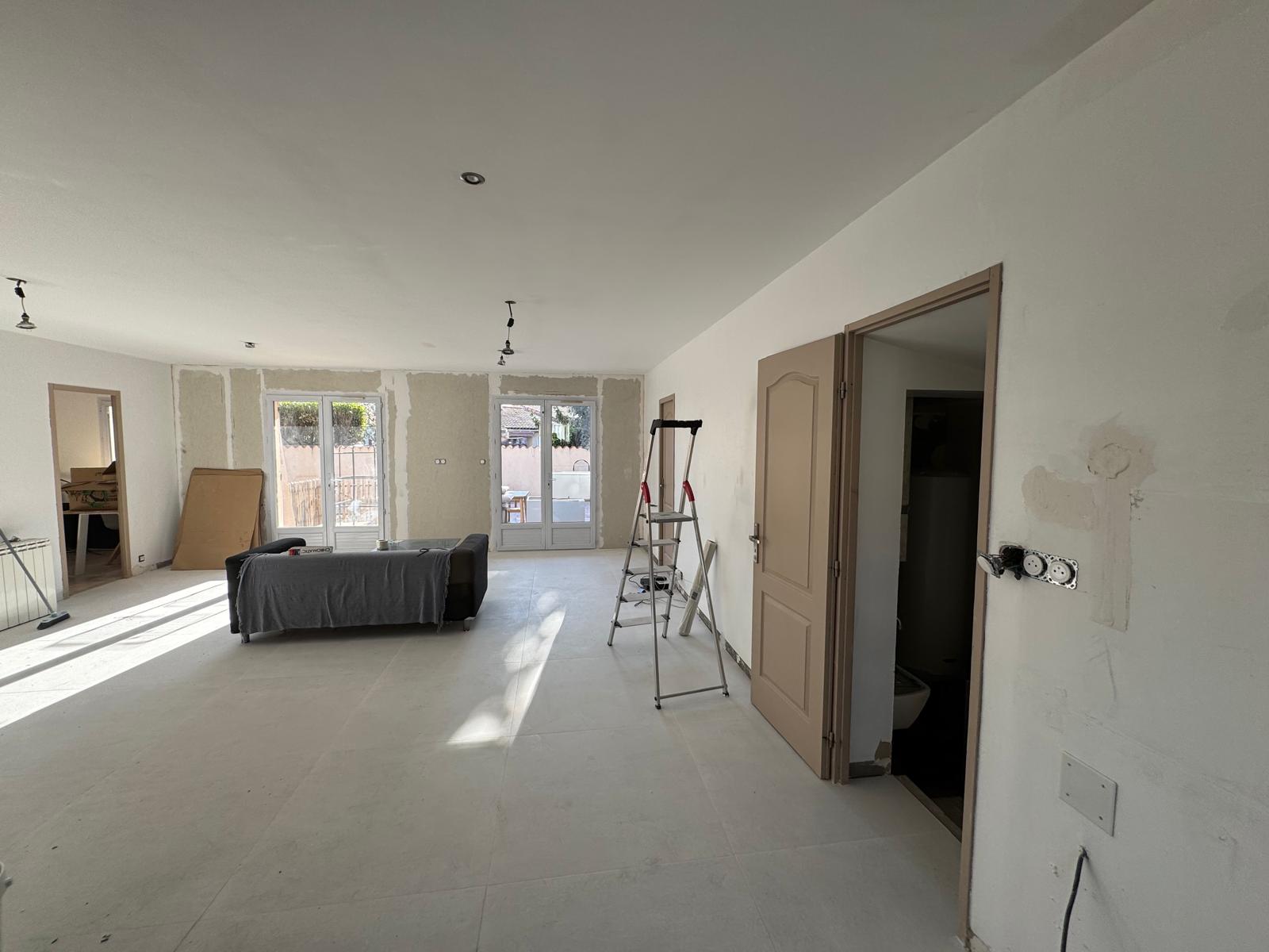 Grand salon en rénovation avec murs blancs, carrelage clair, canapé protégé, escabeau et baies vitrées lumineuses.