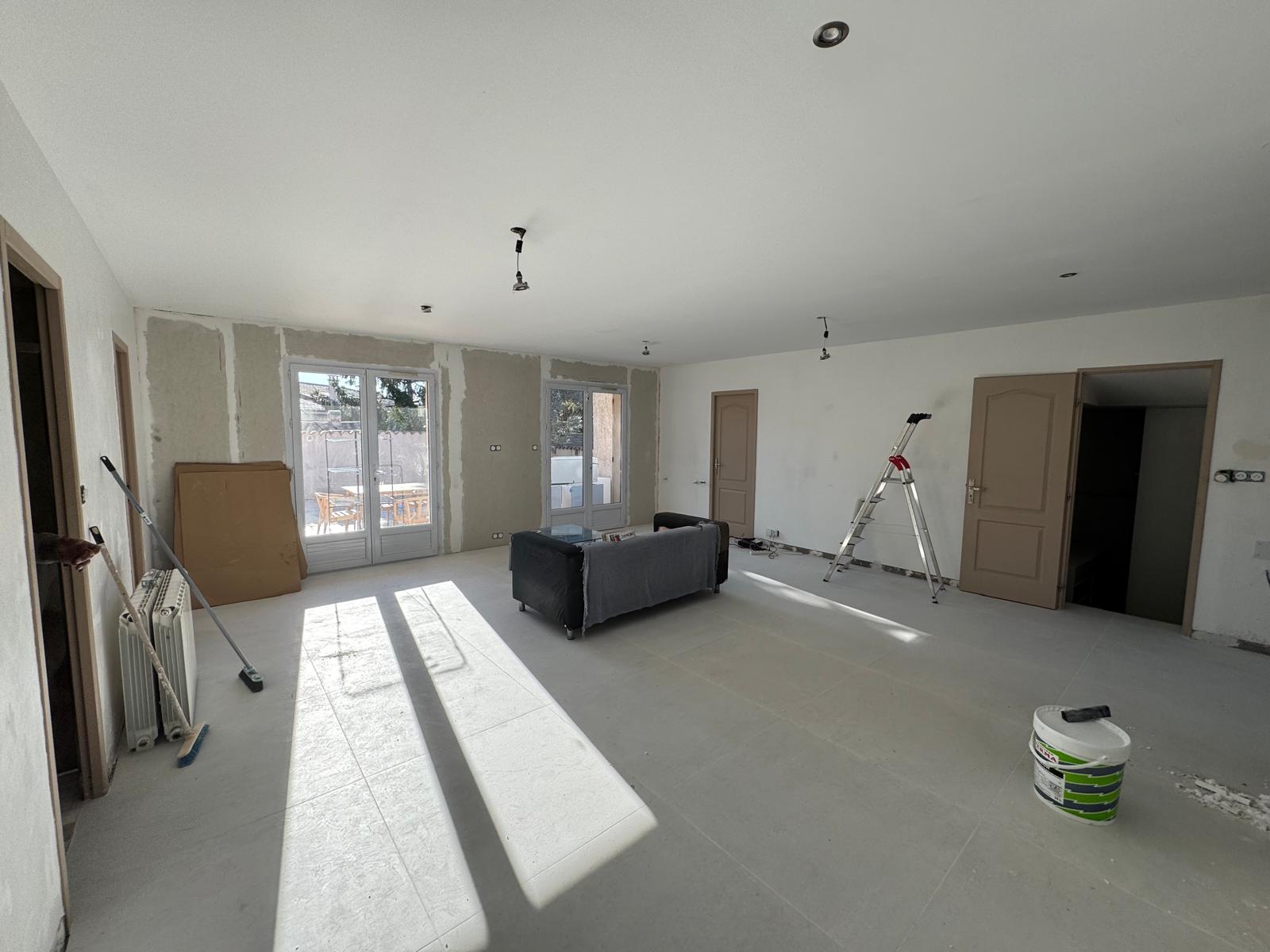 Rénovation d'un salon spacieux avec murs en plâtre, carrelage clair, canapé central, échelle et matériel de peinture.