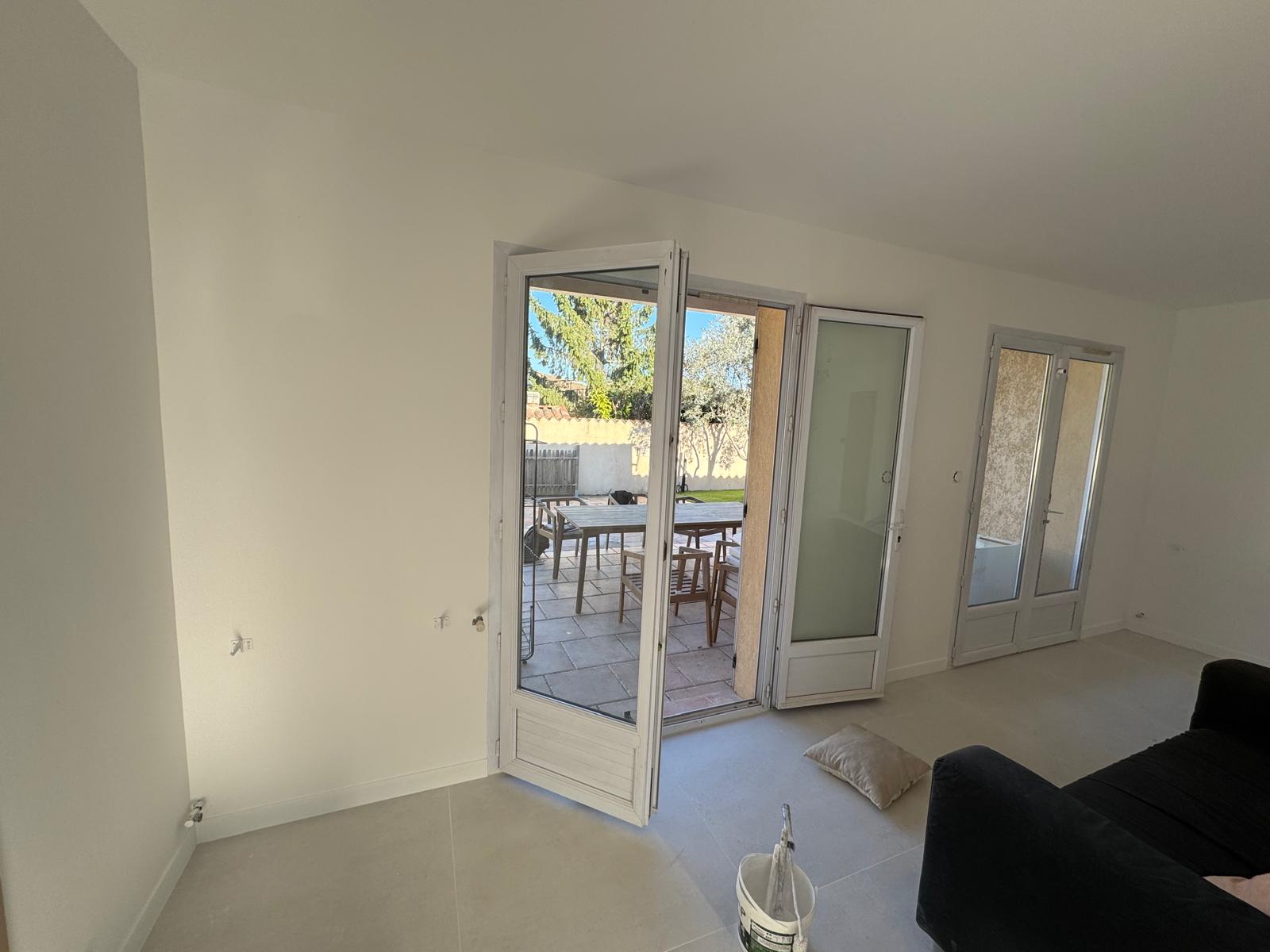 Salon blanc lumineux avec porte-fenêtre ouverte sur terrasse. Rénovation en cours.